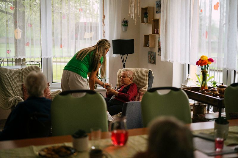 A caregiver crouched to eye level beside an older woman in an armchair