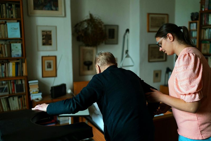 A younger woman steadying an older man at a piano in a book-lined room